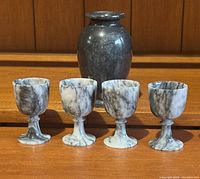 Dark marble vase with rounded body and flared rim next to four small marble shot goblets in white and gray veining, displayed on wooden surface.