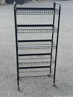 Front view of black metal wire shelving unit with five wire mesh shelves and caster wheels at base against outdoor pavement background.