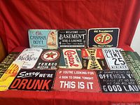Front view of 15 assorted metal tin decorative signs with vintage, automotive, and humorous themes arranged on red fabric background.