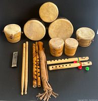 Lot of 13 musical instruments including various traditional hand drums, wooden flutes, a harmonica, and drumsticks arranged on black background.