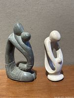 Photo showing both dark gray and light beige soapstone abstract family statues side by side on wooden surface against plain background