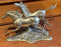 Photo showing side view of two brass horses running sculpture on wooden surface, details of horse musculature and flowing manes visible.