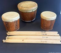Three wooden bongo drums with natural leather skins and nine wooden sticks laid in front, showing general condition and size.