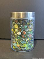 Photo of a clear plastic jar with a silver metal lid filled with colorful vintage glass marbles of various patterns and colors.