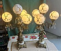 Pair of vintage brass and marble table lamps each with five white frosted globes featuring gold decorative vine patterns and hanging crystals, placed on decorative floral tablecloth.