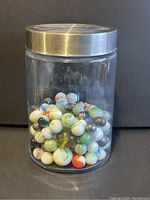 Front view of clear glass jar filled with assorted vintage glass marbles in various sizes and colors including white, green, black, orange, and speckled patterns.