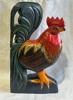 Front and side views of a hand carved and painted wooden rooster figurine, showing detailed feathers, comb, and tail with vibrant coloring.