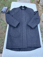 Full view of the charcoal gray men's wool coat laid flat on a table outdoors. Coat has a clean, sleek silhouette with rounded patch pockets and black collar. Shows front exterior and length.