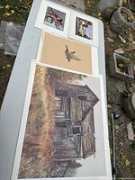 Four artworks laid on a white table outdoors, including a large print of an old barn-like house, a bird flight print, and two smaller images of buildings.