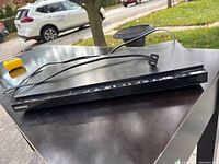 Samsung Blu-ray player unit with attached power cord, showing front and side profile, black color, placed on outdoor surface.