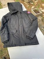 Full view of Canada Goose black men's jacket on table outdoors, showing hood, pockets, buttons, and Canada Goose patch on left sleeve.