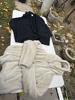 Two items laid flat on a white table outdoors: a black sweater vest and oatmeal fleece zip-up hoodie.