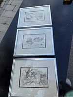 Three silver framed black and white sketches by Jo Ko displayed on a black surface outdoors.