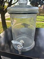 Front and side views of the clear glass beverage dispenser showing the removable glass lid and chrome spigot.