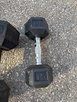 Photo shows two dumbbells on asphalt ground. The 15 pound dumbbell has chrome handle and black hex rubber-coated weights marked '15'.
