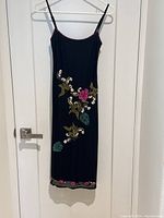 Front view of black fitted dress with colorful floral embroidery, beading and sequins, hanging on white hanger on door.