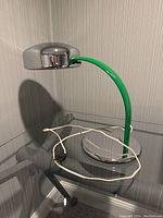 Full view of the lamp sitting on a glass table showing chrome shade, green arc arm, and round base with attached white power cord.