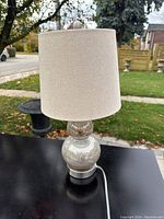 Front view of modern table lamp with linen-look oatmeal lampshade and reflective silver base on black surface outdoors.