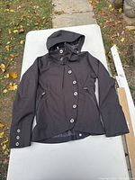 Full view of the black NAU women’s jacket laid flat showing removable hood, button details on front and cuffs