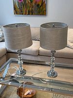 Two modern table lamps on glass table, chrome bases with three clear glass ball stems, oatmeal linen-look fabric shades, power cords visible.