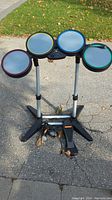 Full front view of the PlayStation Rockband drum set on outdoor pavement, showing four colored drum pads on adjustable stands with foot pedal and microphone cables near the base.