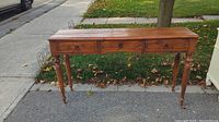 Front view of rustic wood table showing three drawers with brass handles and turned legs, placed outdoors on sidewalk.
