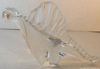 Clear glass dinosaur paperweight seen from a side angle showing the sail back, head, and tail.