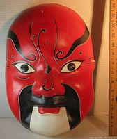 Front view of large red Asian papier mache mask with detailed black and white facial designs showing size in relation to ruler