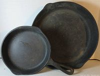 Photo of top and cooking surfaces of a smaller 6.5 inch contemporary cast iron skillet and a larger 10 inch vintage cast iron skillet with raised ring base, both showing wear and use marks.