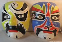 Front view of two colorful papier mache Asian masks placed side by side with detailed painted features.