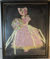 Full view of vintage framed paper doll in pink dress holding bouquet made of fabric flowers with lace and ribbons