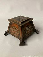 Front angled view of bronze/copper-toned square inkwell with raised silver leaf motif and curled scroll feet.