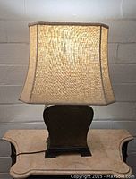 Full view of lit lamp showing beige linen lampshade and brass base with geometric pattern.