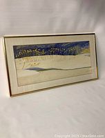 Front angle of framed watercolor showing winter landscape with dried grasses in snow and creek under blue sky.