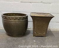 Both planters side by side against a white wall and concrete floor, showing their different shapes and patina.