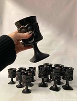 Hand holding one black glass goblet showing size and shape against neutral background. Set of 12 black glass goblets grouped behind.