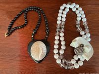 Two large vintage statement necklaces side by side on wooden surface; black beaded necklace with heart-shaped mother-of-pearl pendant and double-strand white and clear bead necklace with geometric rhinestone centerpiece.