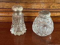Front view of two antique cut glass salt shakers with silver tops, showing detailed glass cuts and silverwork on tops