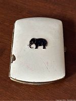 Front view of cigarette case showing black enamel elephant design and surface wear, scratches, and enamel chipping.
