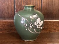Front view of a small dark green cloisonné vase with white floral blossom design and brass rim.