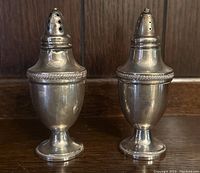 Front close-up view of two sterling silver shakers on wooden surface with wall in background, showing overall condition and shape.