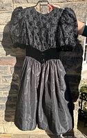 Vintage 1980s black party dress showing puffed sleeves, black sequined bodice, velvet waistband and gathered skirt in sunlight, hanging on hanger.