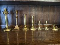 Seven brass candlesticks displayed on a wooden shelf with a dark wood paneled background. Shows the entire collection from left to right.