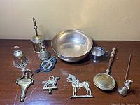 Photo showing all the brass items grouped including bowl, horse brasses, bells, snuffers and door knocker on a wood surface.