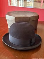 Black Knox superfine wool felt bowler hat with sewn grosgrain ribbon band shown sitting on wooden table, with original octagonal cardboard hat box in background.