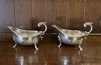 Pair of two vintage silver plated sauce boats shown side by side on wooden surface, decorated with embossed scallop patterns and clawed feet.