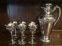 Six silver plated stemmed goblets arranged alongside matching pitcher on wood shelf