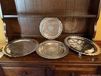 Four vintage silver plated serving pieces displayed on wooden cabinet shelf: a decorative round platter, two large trays, and a Wells meat dish on feet showing some copper through plating.
