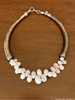 Photo showing full view of the necklace laid flat on a wooden surface, displaying the silver metal band and the pale white, pink, and purple stones in clusters at the front center.