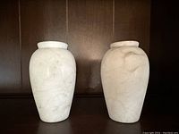 Photo of both alabaster vases side by side on a dark wooden surface against a wood-paneled background. Shows size and shape of both items.